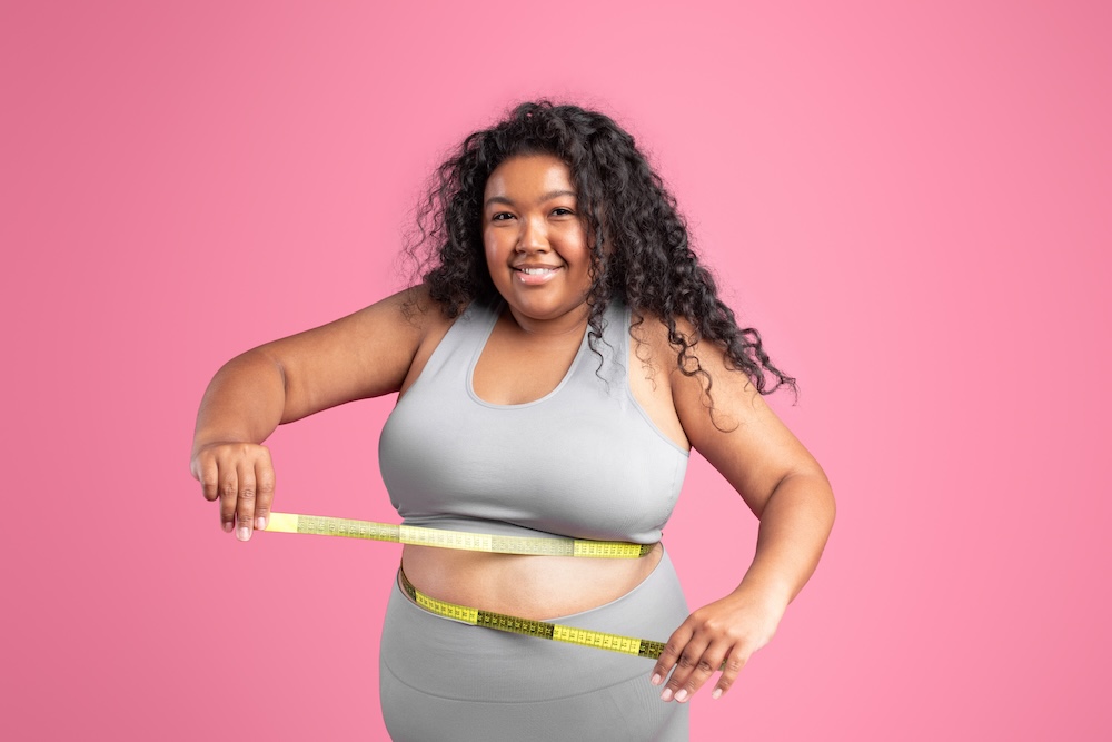 Overweight woman wearing work out clothes smiles and wraps a measuring tape around her stomach to determine how many pounds she would like to shed at the weight loss clinic in Oklahoma City